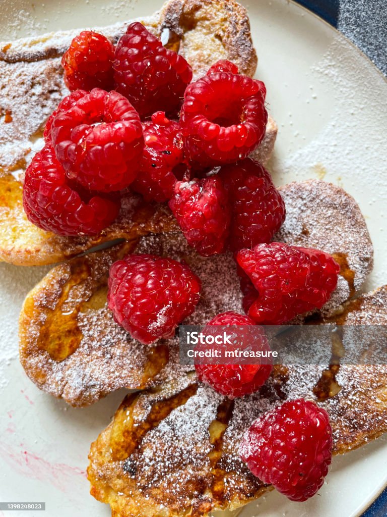 Close-up image of white plate of golden French toast cooking in non-stick frying pan, brioche bun eggy bread topped with raspberries, sprinkled with icing sugar, drizzled with honey, elevated view Stock photo showing close-up, elevated view of brioche, homemade eggy bread often called French toast on white plate. French Toast Stock Photo