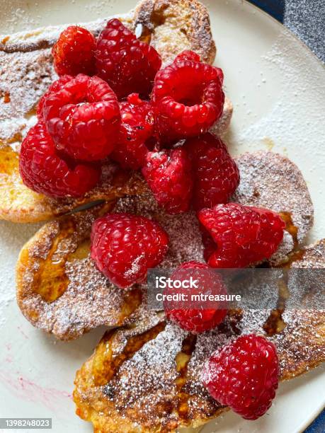 Closeup Image Of White Plate Of Golden French Toast Cooking In Nonstick Frying Pan Brioche Bun Eggy Bread Topped With Raspberries Sprinkled With Icing Sugar Drizzled With Honey Elevated View Stock Photo - Download Image Now