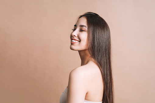 Beauty nude portrait of happy young beautiful asian woman with healthy dark long hair in top bando on beige background