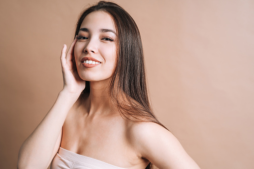 Beauty nude portrait of happy young beautiful asian woman with healthy dark long hair in top bando on beige background