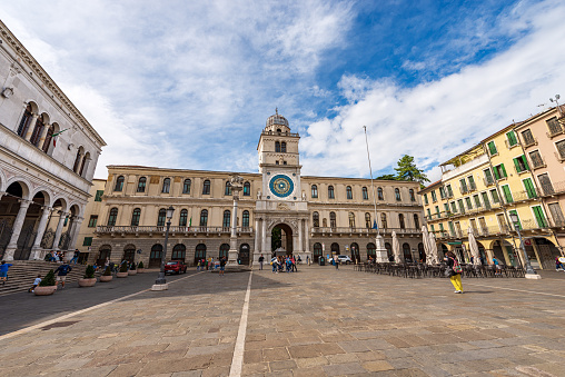 Padua, Italy - September 27, 2020: Piazza dei Signori or Piazza della Signoria, town square in Padua downtown, with the Clock Tower (astronomical clock with the constellations of the zodiac) and the palaces of Capitanio and Camerlenghi; Winged Lion of Saint Mark, symbol of the evangelist, the Venetian Republic and the Veneto Region. Veneto, Italy, Europe. A group of people relax sitting in the bars that surround the square or stroll.