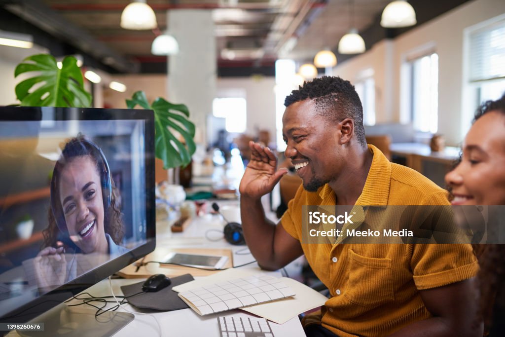 A Black man waves to his colleague on a video call from his office Hybrid Workplace Stock Photo