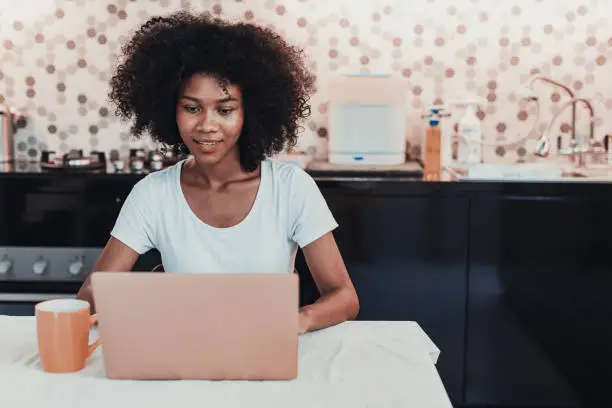 Photo of Young woman working using laptop at home in kitchen.