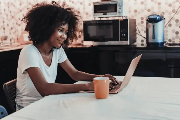 Photo of Young woman working using laptop at home in kitchen.