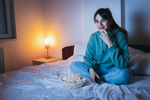 Young woman wearing pajamas sitting in bed, eating popcorn and laughing while watching  funny movie on TV, relaxing at home late at night