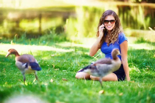 Attractive young woman in her late teens, with long curly brown hair, has a happy conversation on her mobile phone as she relaxes on grass beside a body of water, surrounded by Egyptian geese (which are, technically, ducks).