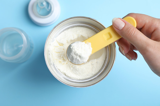 Woman taking powdered infant formula with scoop from can on light blue background, top view