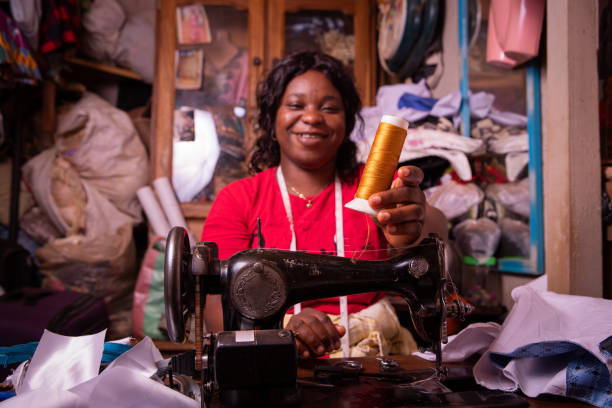 una sarta africana tiene in mano una bobina e lavora nella sua sartoria, è felice e sorridente - manual worker sewing women tailor foto e immagini stock