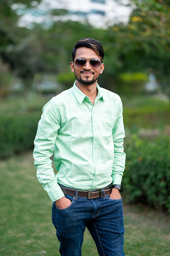 Outdoor portrait image  of a young handsome man of Indian origin with hands in pocket , wearing formal shirt and eyeglasses while standing outside at park in city.