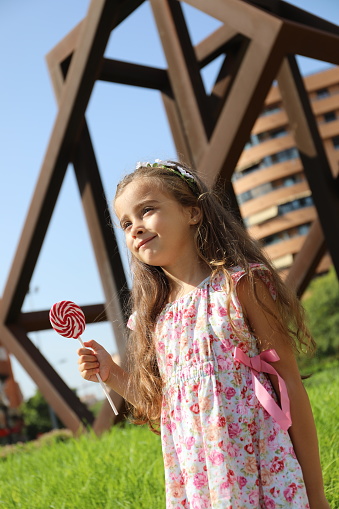 a pretty girl with brown hair and light green eyes, wearing a flowery dress, happy and smiling with a lollipop in the city of alicante