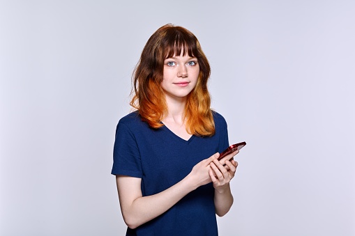 Serious sad teenage female with a smartphone in her hands looking at the camera on a light studio background