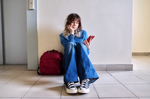 Sad young female student with backpack smartphone sitting near elevator on floor. Lonely frustrated teenage female, frustration problem pain depression concept