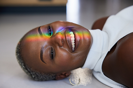 Close-up of a ray of rainbow colored light refracting onto a the face of a woman lying on a floor and smiling