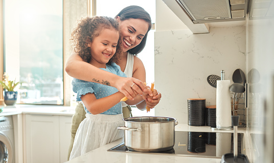 Mother and little daughter cooking together in the kitchen. Mixed race mother and child standing by the stove breaking spaghetti and throwing it in boiling water