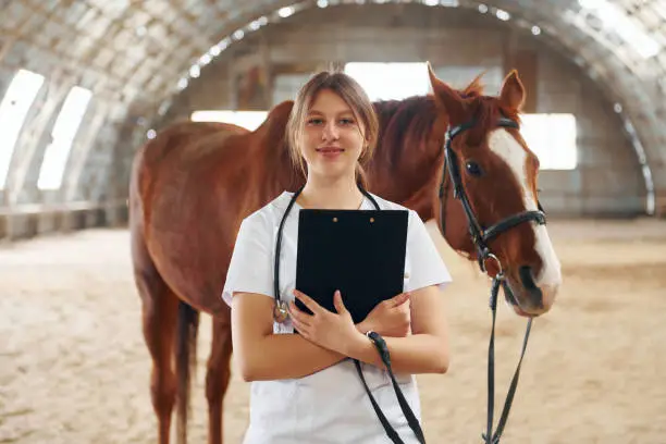 Holding notepad in hands. Female doctor in white coat is with horse on a stable.