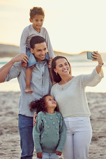 Happy cheerful mixed race family smiling for a selfie spending time at the beach together. Hispanic mother smiling taking a photo with her children and husband with her cellphone bonding on vacation