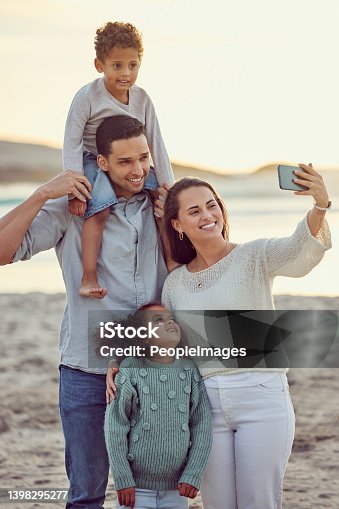 istock Happy cheerful mixed race family smiling for a selfie spending time at the beach together. Hispanic mother smiling taking a photo with her children and husband with her cellphone bonding on vacation 1398295277