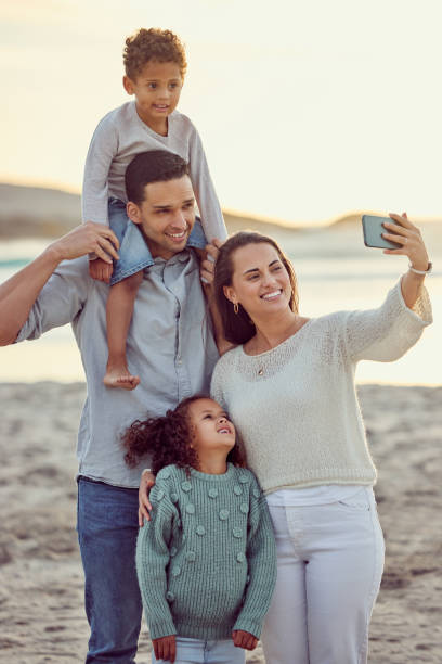 счастливая веселая смешанная семья улыбается для селфи, проводя время на пляже вместе. испаноязычная мать улыбается, фотографируясь со сво - vertical photography стоковые фото и изображения