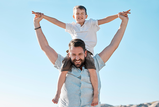 A young boy with outstretched arms sits on the father's shoulders and imagine a flight. Outdoors portrait of cheerful father with son.
