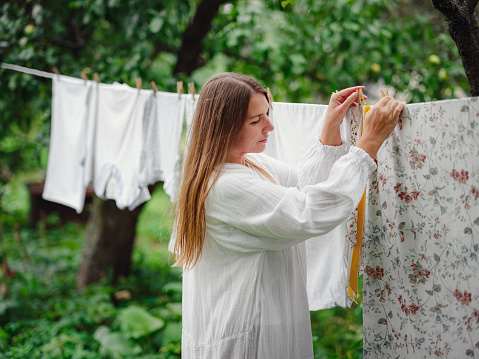 middle aged caucasian woman in white dress doing homework, hanging clothes on clothesline on street in courtyard of village cottage house, laundry day concept