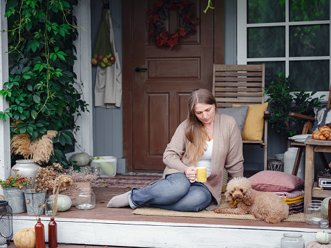 middle aged caucasian woman in brown cardigan sitting on terrace of house with her beige poodle and enjoying warm autumn weather and drinking morning coffee, autumn lifestyle concept