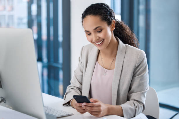 istock Young mixed race female using a cell phone and sending a text while sitting in front of her computer in a office. Hispanic businesswoman using a wireless device while browsing the web 1398313393