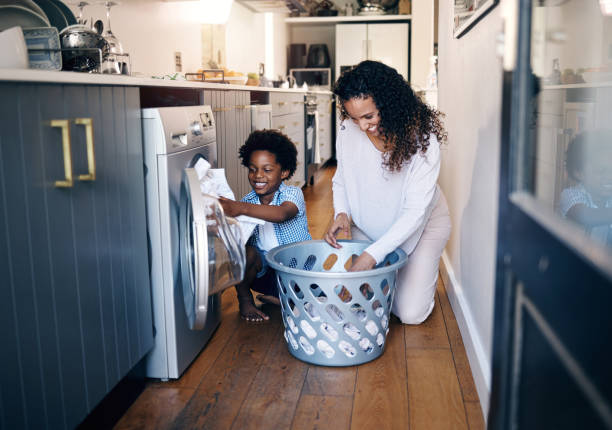 adorable petit garçon afro-américain avec un afro souriant et s’amusant tout en faisant le ménage avec sa mère à la maison. plan métis d’un enfant mignon pliant du linge avec sa mère - tâches ménagères photos et images de collection