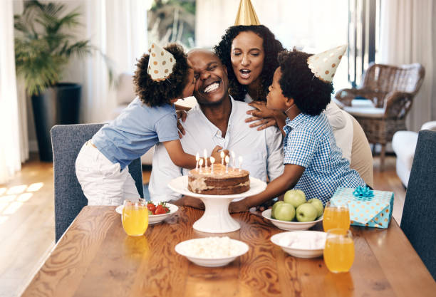 afroamerikanische familie trägt partyhüte und feiert einen geburtstag zu hause mit kuchen. kleine geschwister umarmen und küssen ihren vater an seinem geburtstag. glücklicher mann lächelt nach einer geburtstagsüberraschung - african ethnicity brother ethnic little boys stock-fotos und bilder