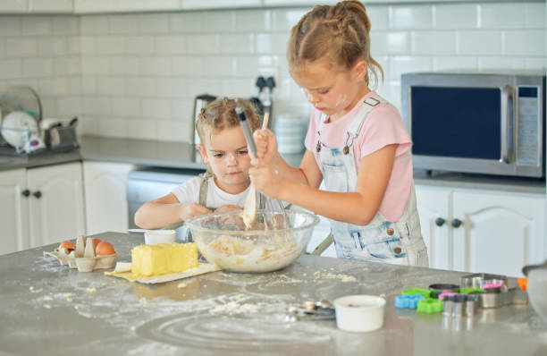 duas garotinhas confusas assando na cozinha em casa. irmãs brancas focadas aprendendo a fazer uma sobremesa e cozinhar. crianças adoráveis se unindo e ajudando uns aos outros para preparar uma receita para a casa - vitality innocence clothing human age - fotografias e filmes do acervo