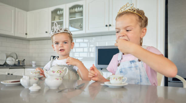 家でお茶会をしている二人の小さな女の子。きょうだいの姉妹の友達がティアラを着てティーセットで遊んだり、キッチンのテーブルでクッキーを食べたり。仲良くなり、一緒に遊ぶ姉妹た� - tea party party tea little girls ストックフォトと画像