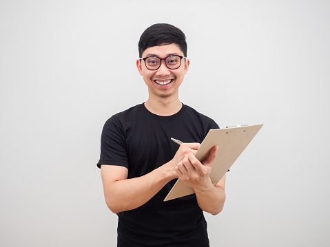 Asian man wearing glasses cheerful smile face writing at document board in hand looking at camera on white background isolated