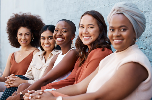 Group of diverse happy businesswomen sitting in a line with coworkers against a wall outside in the city. Businesspeople smiling while sitting outdoors with colleagues