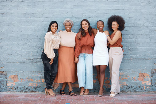 Grupo de cinco jóvenes mujeres de negocios alegres y felices abrazadas mientras están de pie contra una pared afuera en la ciudad. Retrato de colegas felices tomados de la mano y sonrientes de pie en una fila al aire libre photo