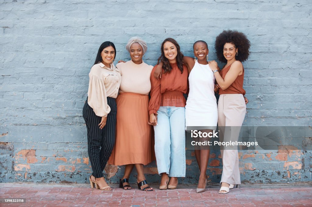 Group of five young happy cheerful businesswomen hugging while standing against a wall outside in the city. Portrait of happy colleagues holding hands and smiling standing in a line outdoors Women Stock Photo