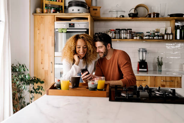 Couple having breakfast in kitchen. Stock photo of a middle aged woman and man having breakfast in a kitchen and laughing together. They are relaxed and looking at a smartphone. two parents stock pictures, royalty-free photos & images