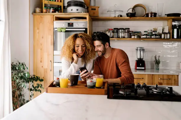 Stock photo of a middle aged woman and man having breakfast in a kitchen and laughing together. They are relaxed and looking at a smartphone.