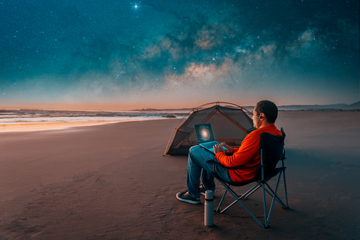 person sitting with laptop on the beach working beside tent under the starry night and milky way