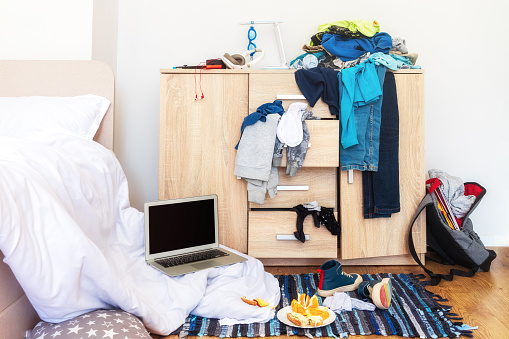 a general view of a teenager's room with a lot of clutter. an open chest of drawers from which things fall out, an unmade bed, food and a laptop on the floor. the concept of raising a teenager