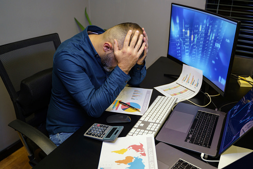 Portrait of stressed male broker sitting with head in hands at desk while working late at his office