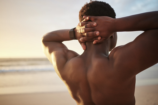 Back of a fit muscle man with hands behind his head, at the beach looking at the view during sunset. Rear view of a fitness guy thinking or bodybuilder staring at the open sky and stretching