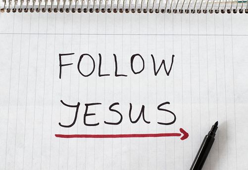 Follow Jesus Christ, handwritten text message with a red arrow showing a way written on a spiral notebook. The biblical concept of the Christian way of life, guidance, and obedience to God. Top view.