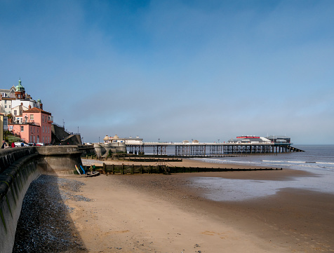 The pier and beach in Cromer, Norfolk, Eastern England, on a sunny day in late March. A few people are walking along the promenade or preparing to go surf boarding; sea mist lurks on the horizon, ready to change the scene within minutes. Cromer Pier is famous for the lifeboat station at the end and for its Pavilion Theatre.