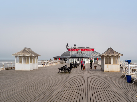 Two people walking dogs along the pier in Cromer, Norfolk, Eastern England, on a day of intermittent sea mist and sunshine. Cromer Pier is famous for its Pavilion Theatre and the lifeboat station at the far end. The town is a typically English, old-fashioned, holiday resort.