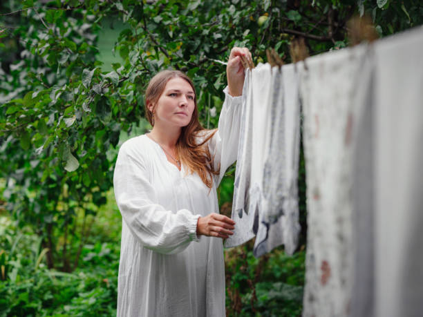 middle aged caucasian woman in white dress doing homework, hanging clothes on clothesline on street in courtyard of village cottage house, laundry day concept