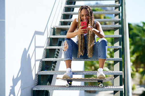 Young black woman with coloured braids, sitting on some steps while consulting her smartphone with her feet resting on a skateboard.