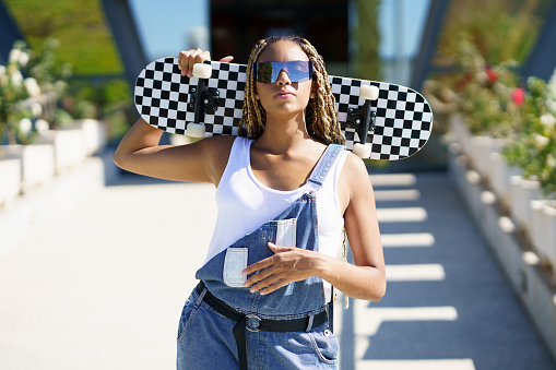 Confident African American female hipster in sunglasses and overalls standing with skateboard in street in summer and looking at camera