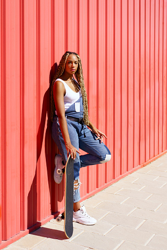 Black young female dressed casual, wtih a skateboard on red urban wall background.