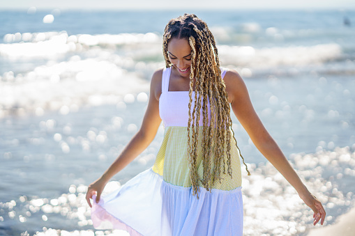 Black girl walking along the shore of the beach wearing a beautiful long dress. Young girl enjoying her vacations.
