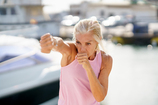 Older woman doing sport in a coastal port. Arm training throwing boxing punches. Mature Female doing shadow boxing outdoors.