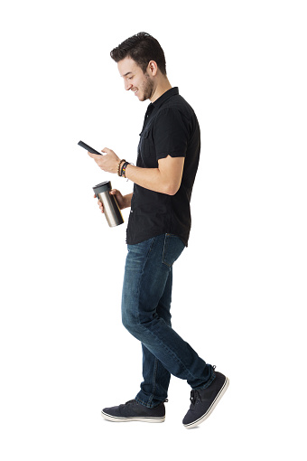 A young latin man walking with a coffee mug in his hand and using a mobile phone.